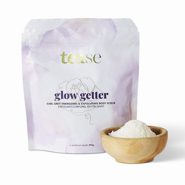 Tease Wellness Glow Getter Earl Grey Body Scrub in pastel purple pouch with wooden bowl of scrub powder.