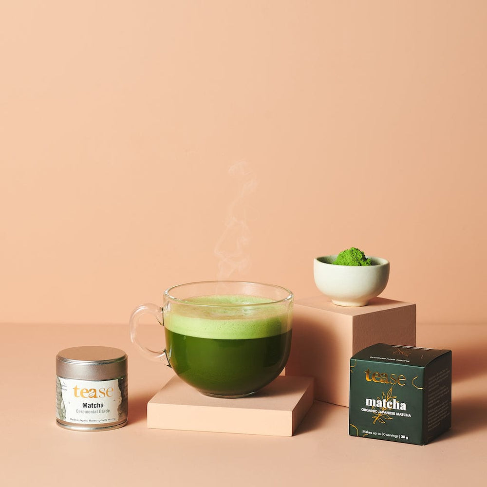 Tease Wellness Organic Ceremonial Matcha in white tin and dark green box with steaming glass cup of vibrant green matcha.