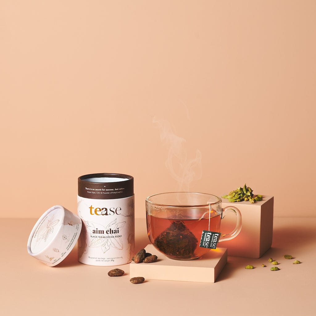 Tease Wellness Aim Chai tea blend in white tube with black lid, steaming chai tea in glass cup, loose spices.