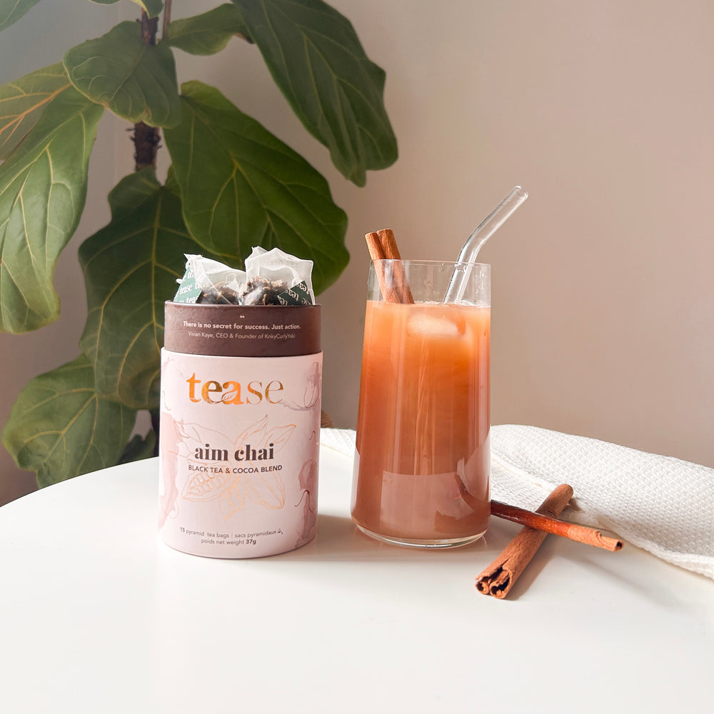 Tease Wellness Aim Chai tea blend in pink cylindrical tube with cinnamon sticks, glass of chai tea with cinnamon stick and.