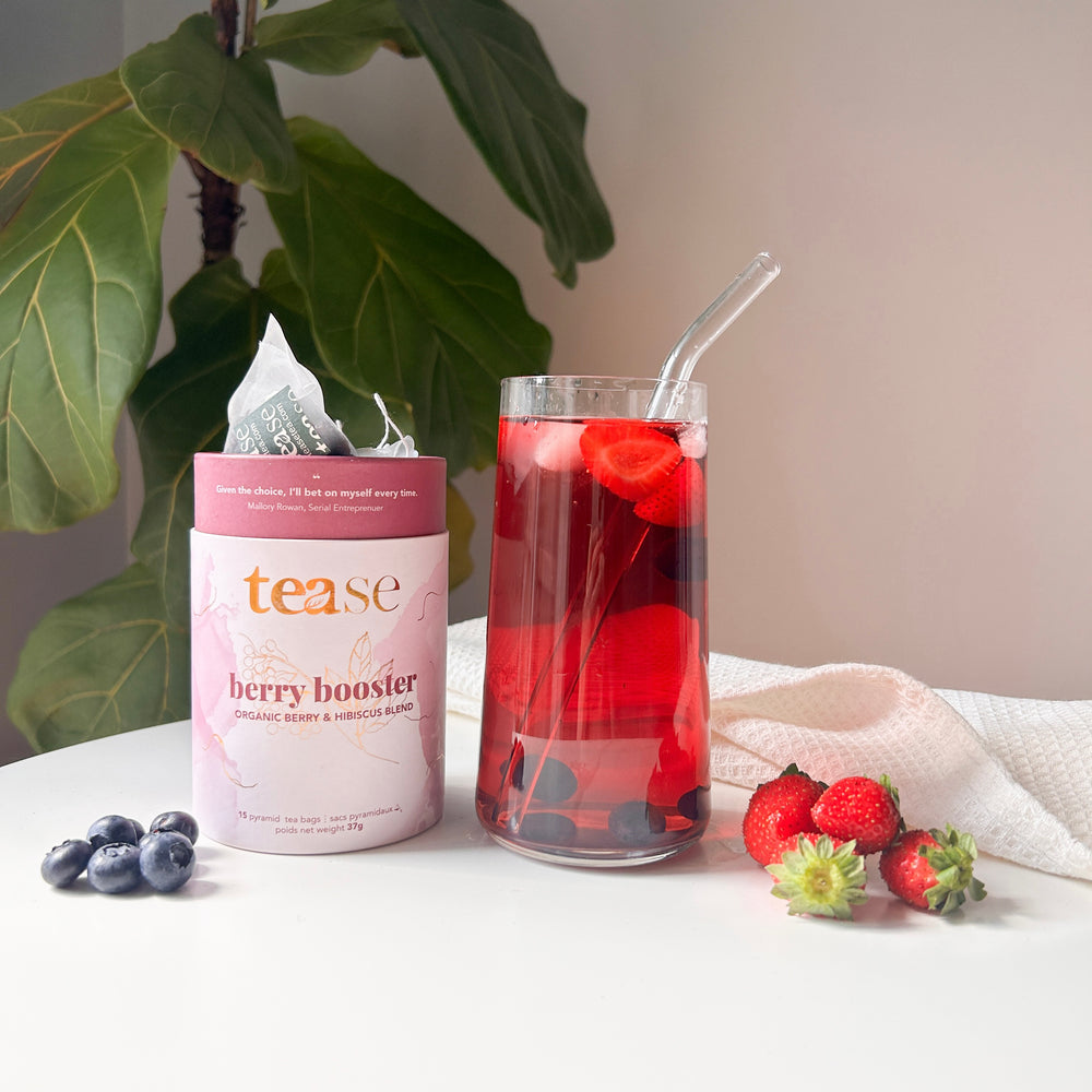 Tease Wellness Berry Booster organic caffeine-free tea blend in pink tube with glass of red berry infusion and fresh berries.