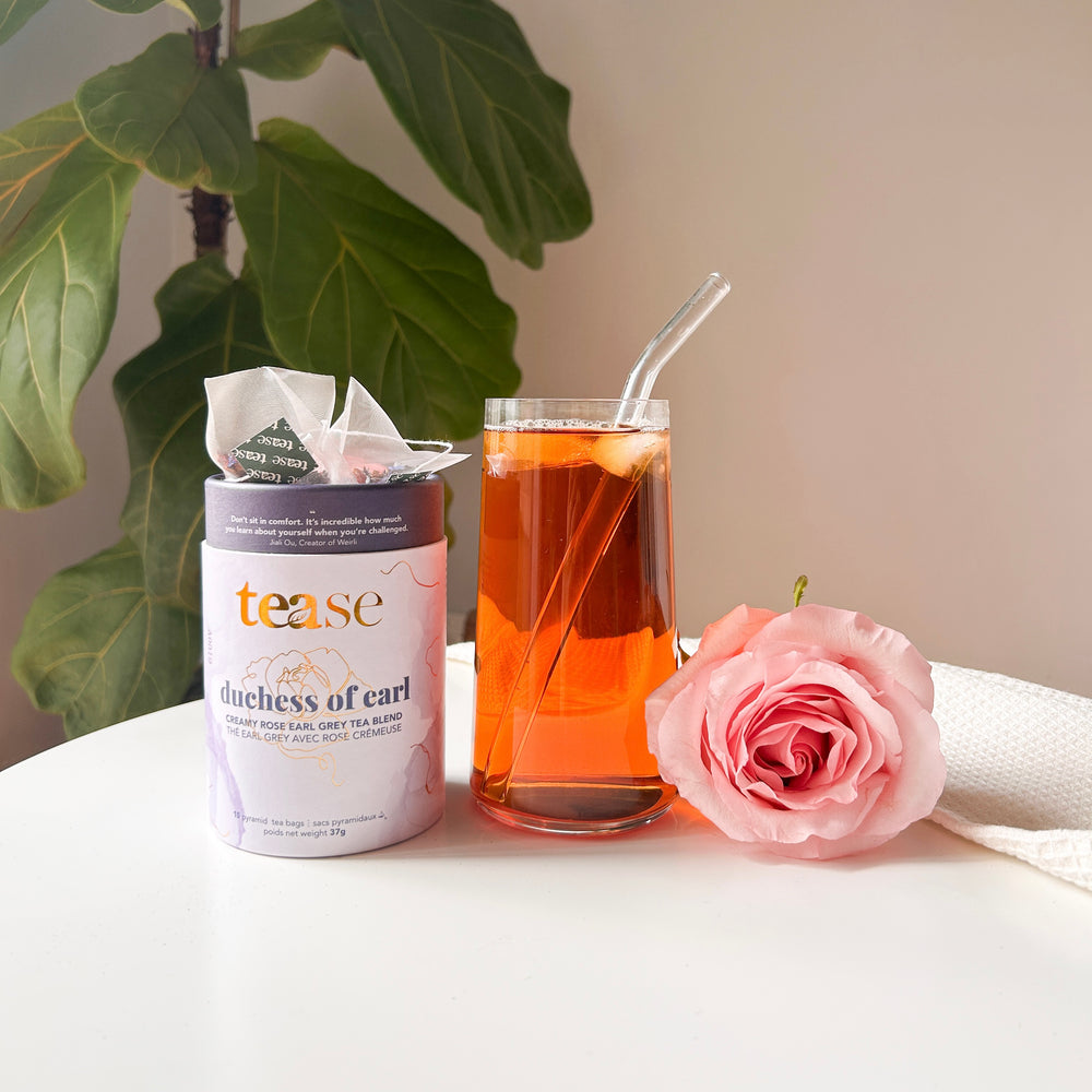 Tease Wellness Duchess of Earl tea blend in lavender tube with glass of amber tea and pink rose on white surface.
