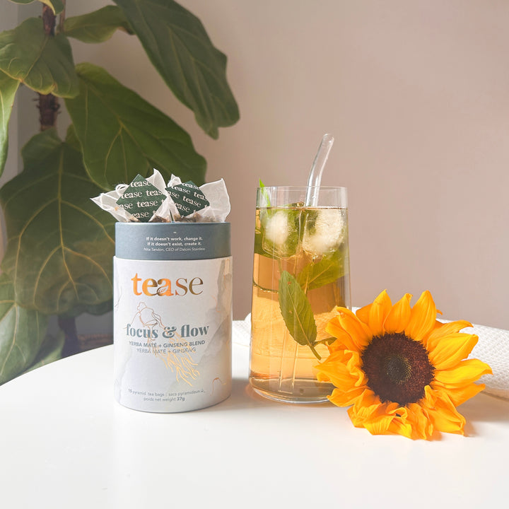 Tease Wellness Focus & Flow tea blend in white cylindrical tube with green tea bag and glass of iced tea garnished with mint .