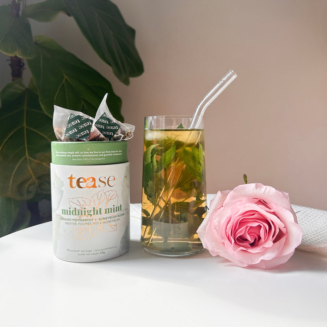 Tease Wellness Midnight Mint organic tea blend in green and white tube with glass of mint tea and pink rose on white surface.