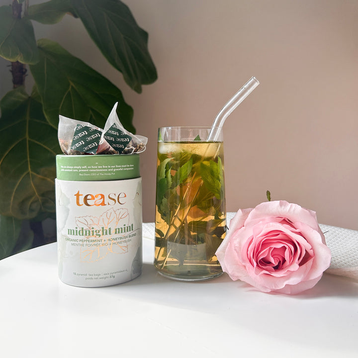 Tease Wellness Midnight Mint organic tea blend in green and white tube with glass of mint tea and pink rose on white surface.