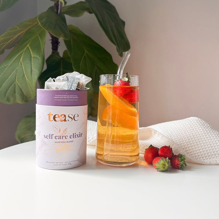 Tease Wellness Self Care Elixir tea blend in purple tube packaging with glass of infused tea and fresh strawberries.