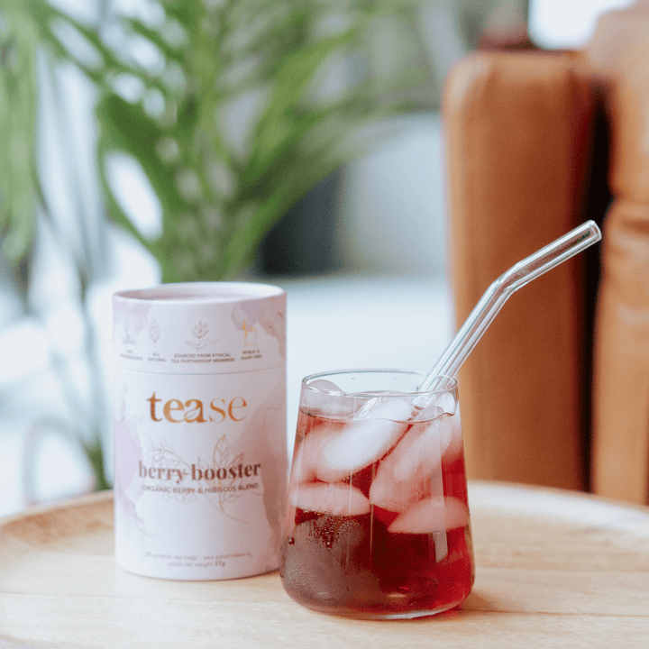 Tease Wellness Berry Booster organic tea blend in white cylindrical tube with glass of iced berry tea and glass straw.