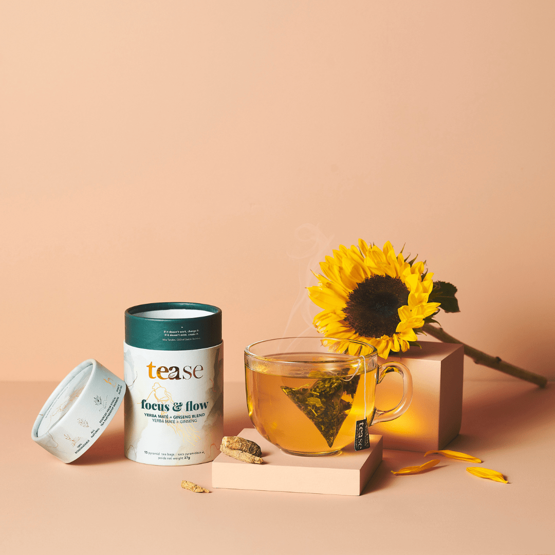Tease Wellness Focus & Flow tea blend in white tube with green lid beside glass cup of brewed tea and sunflower on peach.