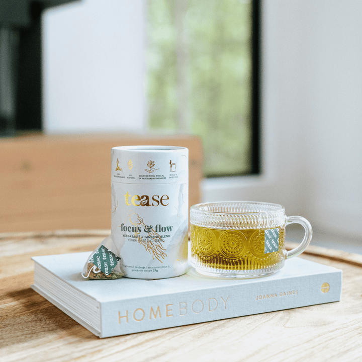 Tease Wellness Focus & Flow tea blend in white cylindrical packaging with herbs illustration, glass cup of brewed yellow tea .