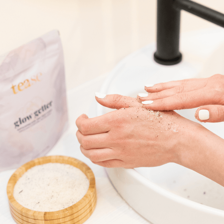 Tease Wellness Glow Getter Earl Grey Body Scrub in white pouch with wooden bowl, showing exfoliating scrub on hands by sink.