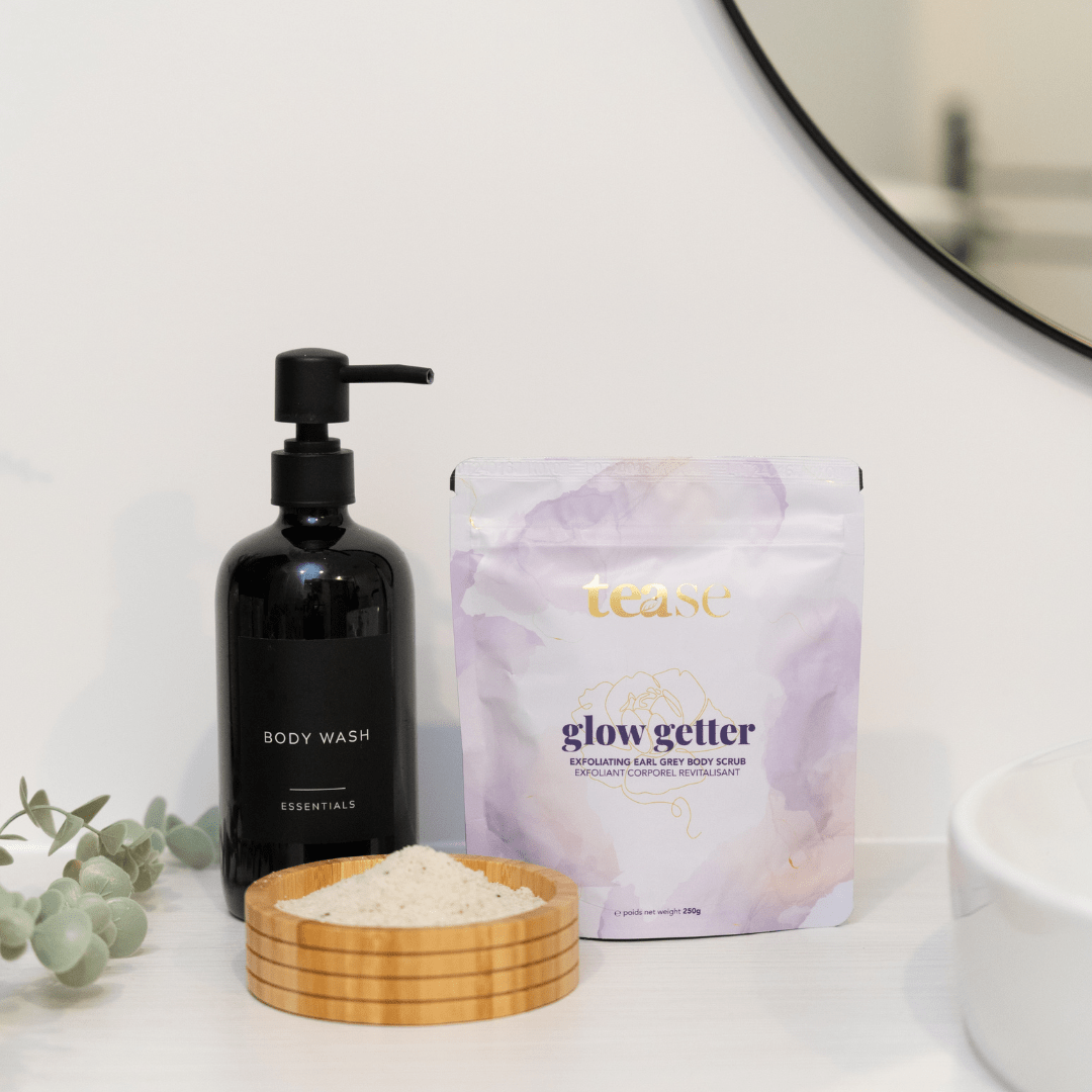Tease Wellness Glow Getter Earl Grey Body Scrub in lavender and white pouch with wooden bowl of scrub powder and black pump.