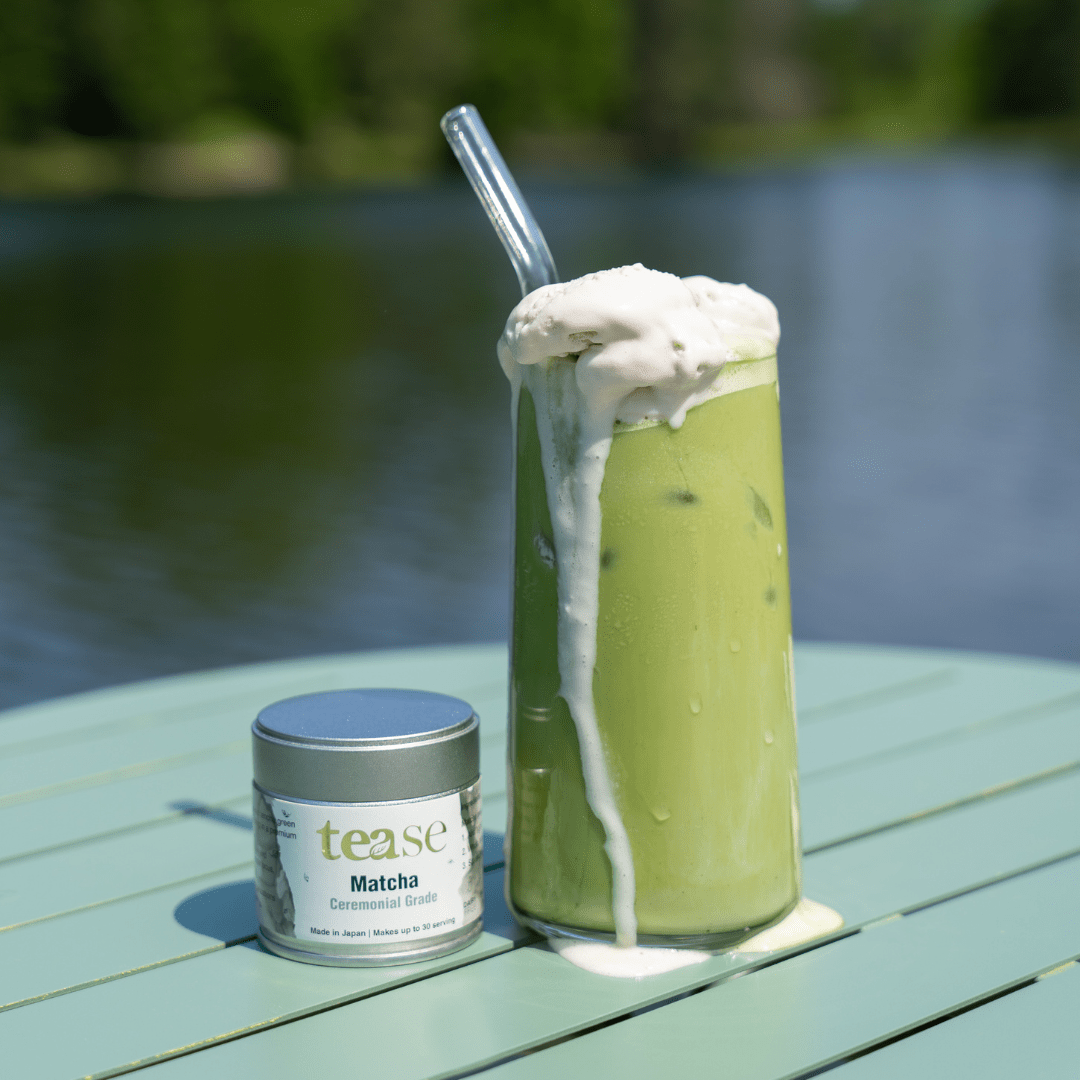 Tease Wellness Organic Ceremonial Matcha powder canister next to tall glass of iced frothy green matcha latte with glass.