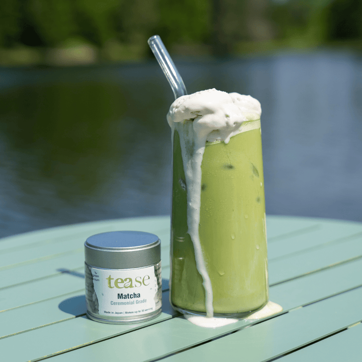 Tease Wellness Organic Ceremonial Matcha powder canister next to tall glass of iced frothy green matcha latte with glass.