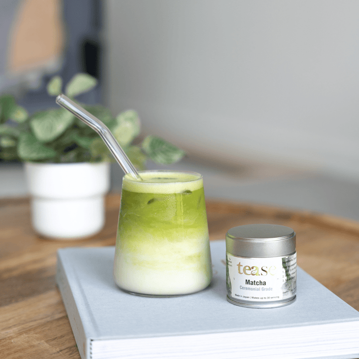 Tease Wellness Organic Ceremonial Matcha in silver tin with green matcha latte in glass and metal straw on book.