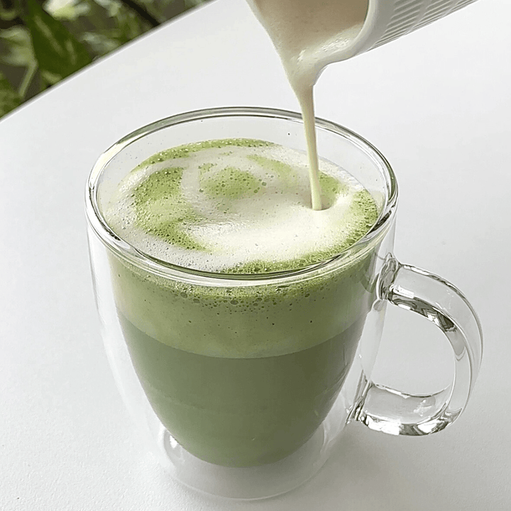 Tease Wellness Organic Ceremonial Matcha frothy green tea in clear glass mug with milk pouring on top.