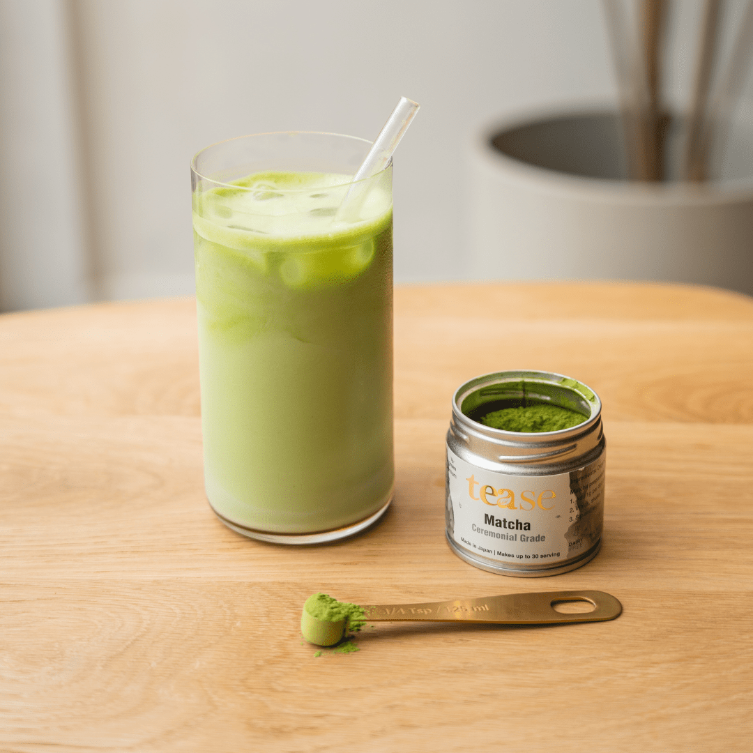 Tease Wellness Organic Ceremonial Matcha tin with green matcha powder on wooden scoop and iced matcha latte in glass with.
