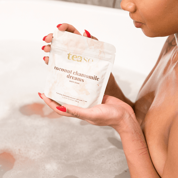 Tease Wellness Coconut Chamomile Dreams bath soak in white resealable pouch held above bubbly bathwater.