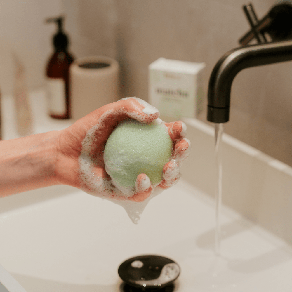 Tease Wellness Matcha Konjac Facial Sponge in hand with foam by a bathroom sink.