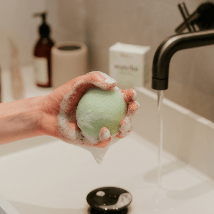 Tease Wellness Matcha Konjac Facial Sponge in hand with foam by a bathroom sink.