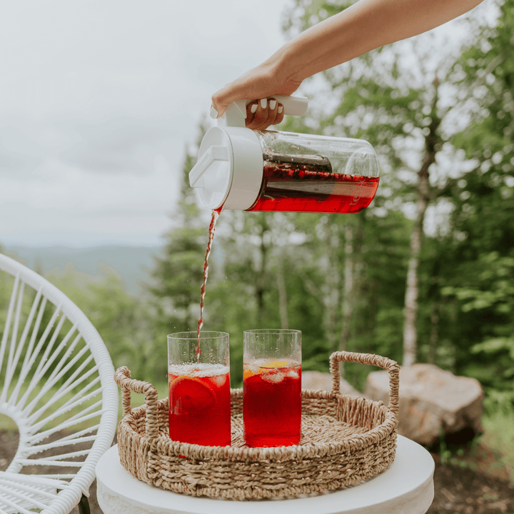 Tease Wellness Cold Brew Iced Tea & Coffee Maker Kit with vibrant red tea pouring into clear glasses on woven tray outdoors.