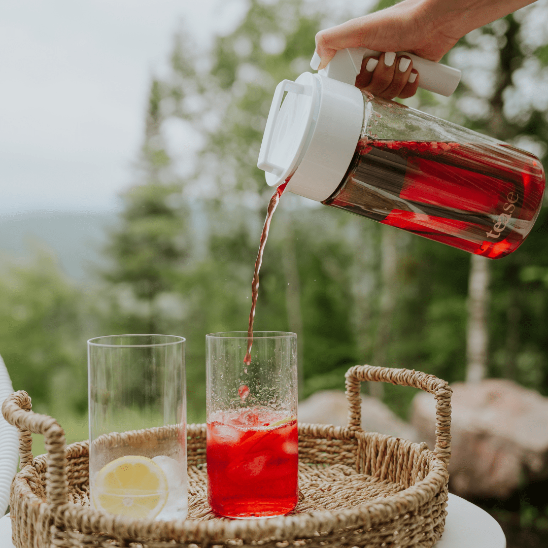Tease Wellness Cold Brew Iced Tea & Coffee Maker Kit pouring red hibiscus tea into glass with lemon slice on tray outdoors.