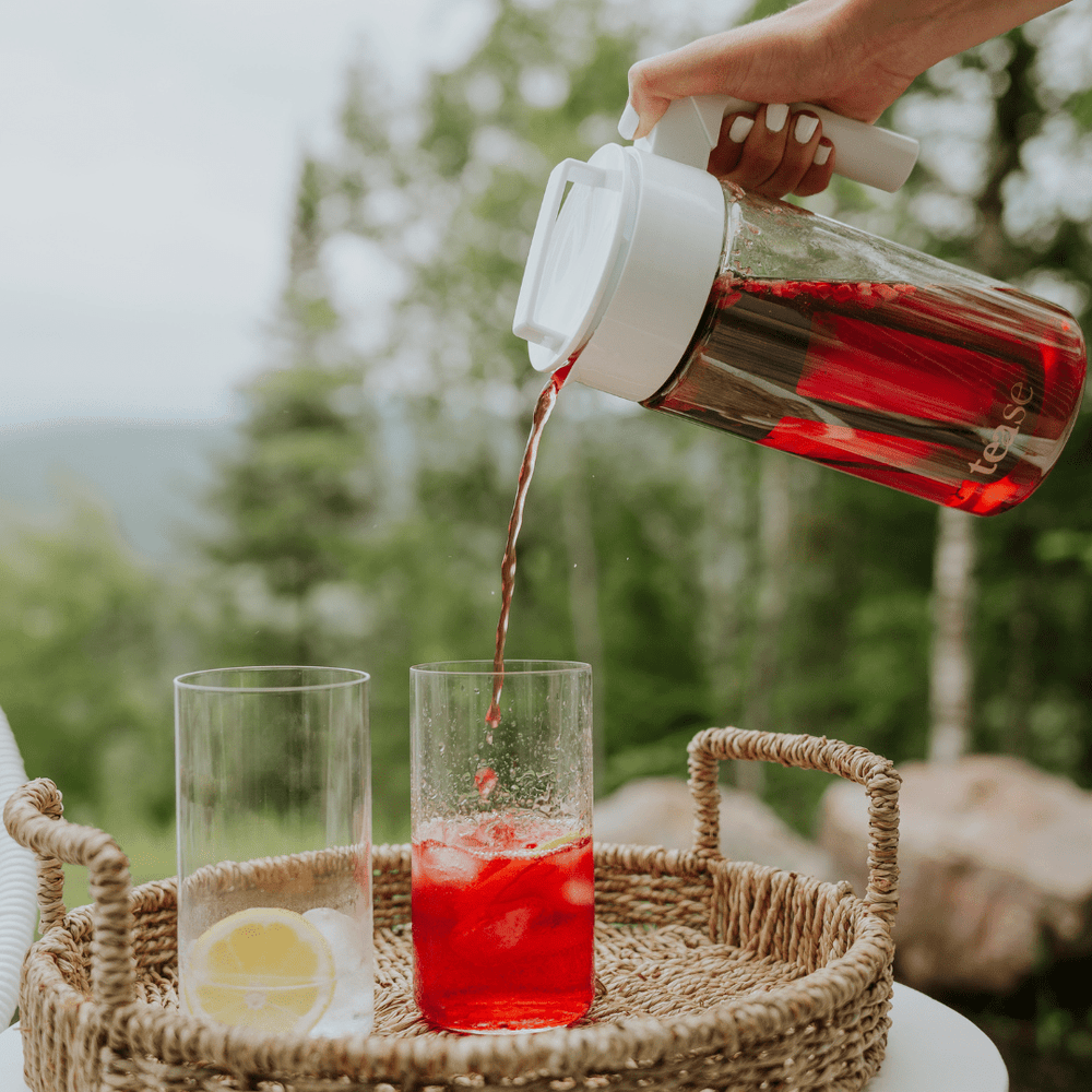 Tease Wellness Cold Brew Iced Tea & Coffee Maker Kit pouring red hibiscus tea into glass with lemon slice on tray outdoors.