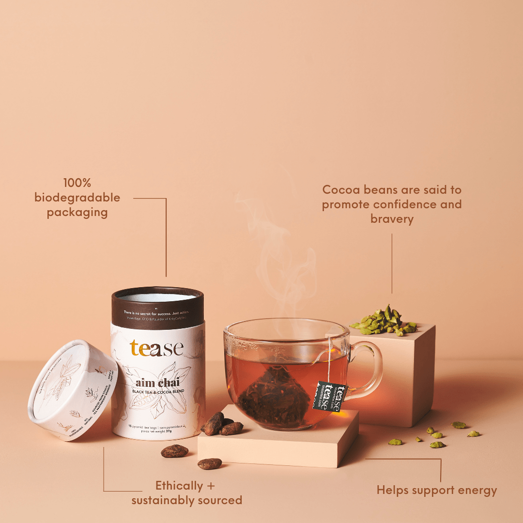 Tease Wellness Aim Chai tea blend in biodegradable tube, with glass cup of chai and cocoa bean tea bag on peach background.