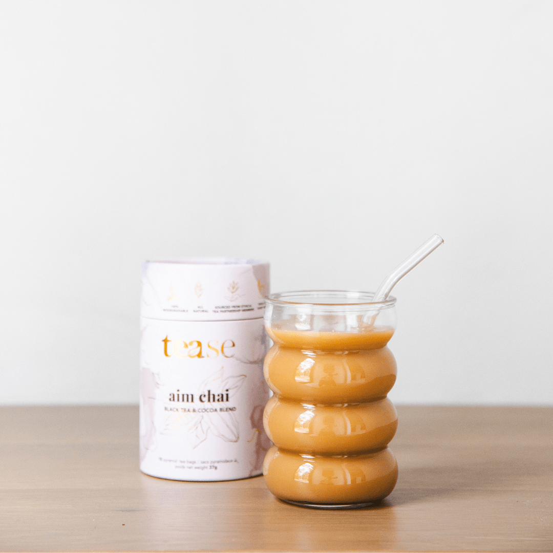 Tease Wellness Aim Chai tea blend in white tube packaging next to glass of frothy chai latte with glass straw.