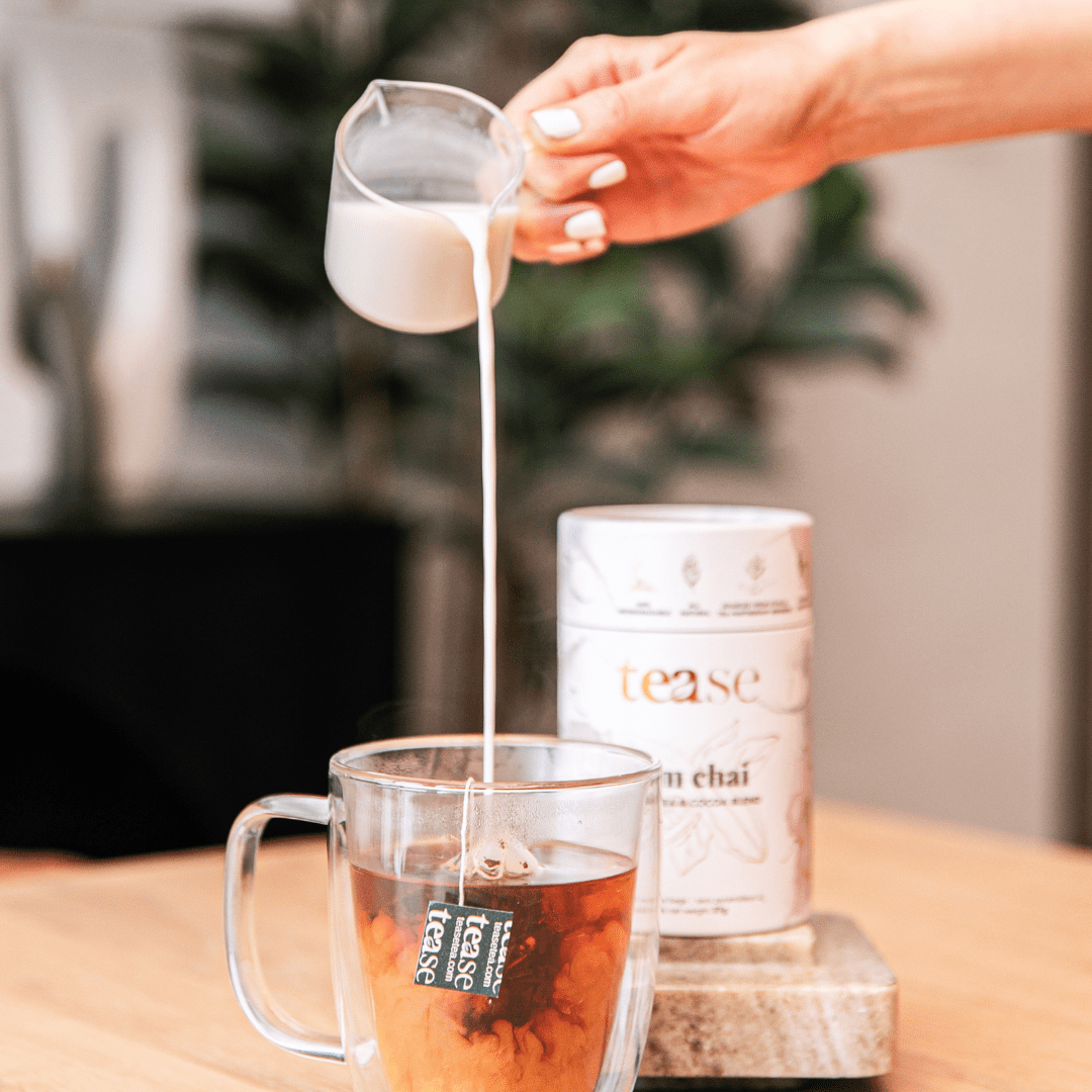 Tease Wellness Aim Chai tea blend in white cylindrical tube with brewed tea in glass cup and milk being poured.