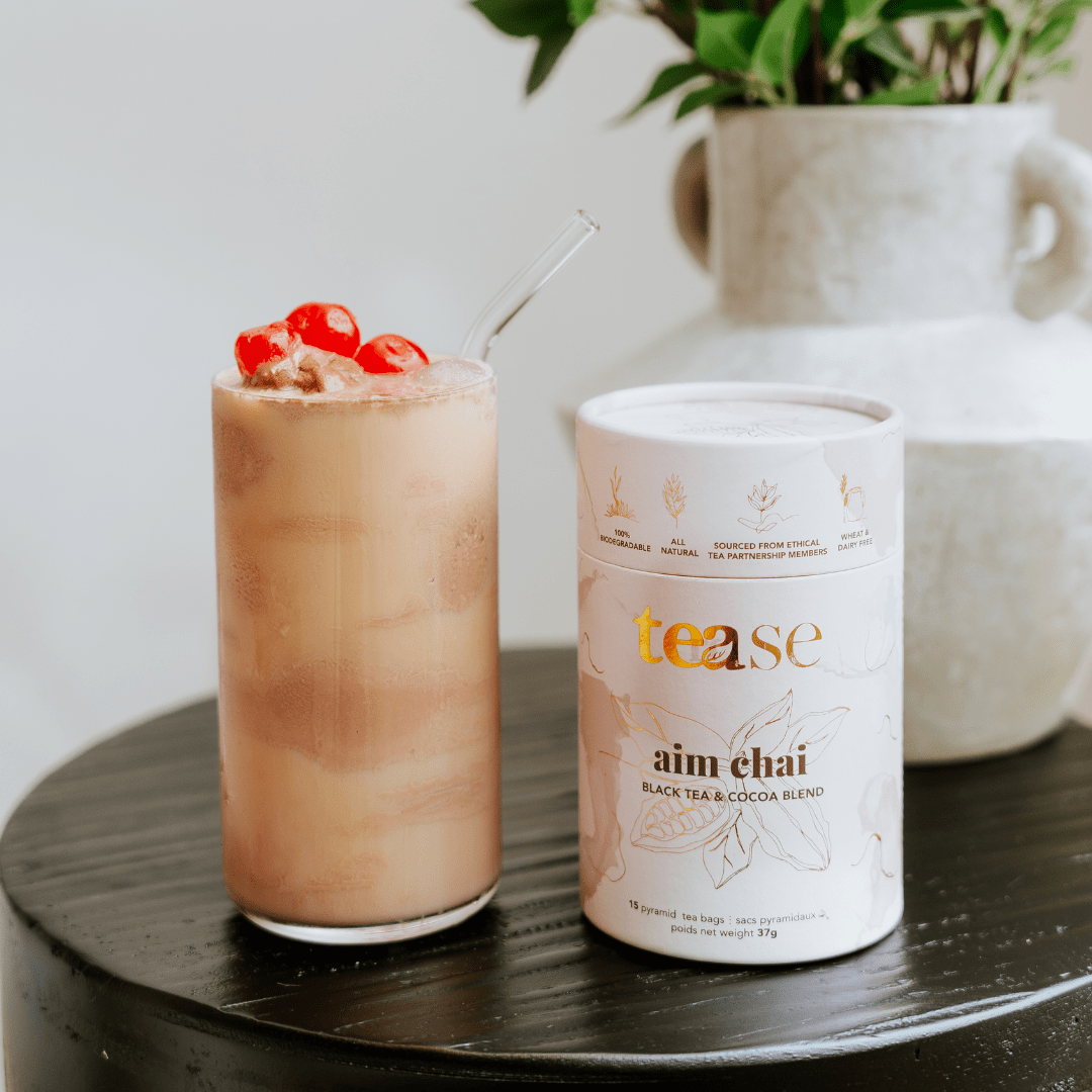 Tease Wellness Aim Chai black turmeric root tea blend in white tube packaging beside iced chai latte with red berries and.