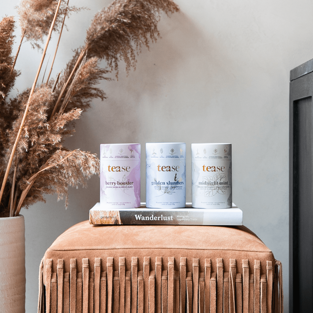 Tease Wellness Organics Gift Set with three tea blends in pastel purple, blue, and white cans on a brown ottoman.